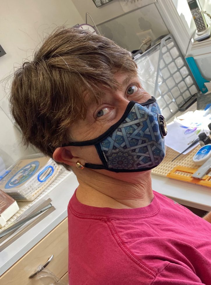 A photo of artist Patty Taliaferro, standing in a glass art studio, wearing a blue respiratory face mask.